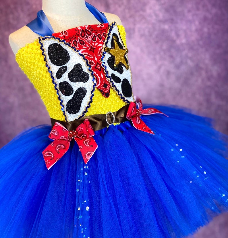 Puede incluir: Un tut&uacute; de tul azul con un corpi&ntilde;o amarillo y rojo con estampado de vaca, un pa&ntilde;uelo rojo y una estrella de sheriff. El corpi&ntilde;o est&aacute; decorado con purpurina.