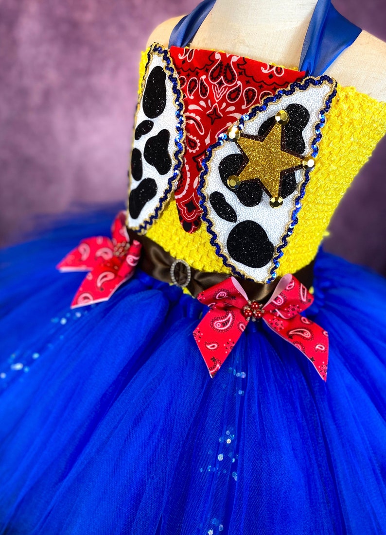 Puede incluir: Un tut&uacute; de tul azul con lazos de bandana rojo y blanco. La parte superior del tut&uacute; es un top de crochet amarillo con un bandana rojo, una impresi&oacute;n de vaca en blanco y negro y una estrella dorada.