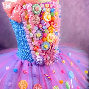 May include: A pink and blue tulle tutu dress decorated with colorful candy, lollipops, and a pink bow. The bodice is blue with a candy design and a pink bow.