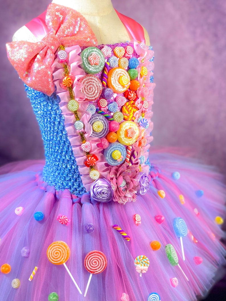 May include: A pink and blue tulle tutu dress decorated with colorful candy, lollipops, and a pink bow. The bodice is blue with a candy design and a pink bow.