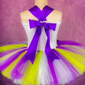 May include: A white tulle tutu skirt with purple, white, and yellow stripes. The skirt has a purple satin ribbon tied in a bow at the back.