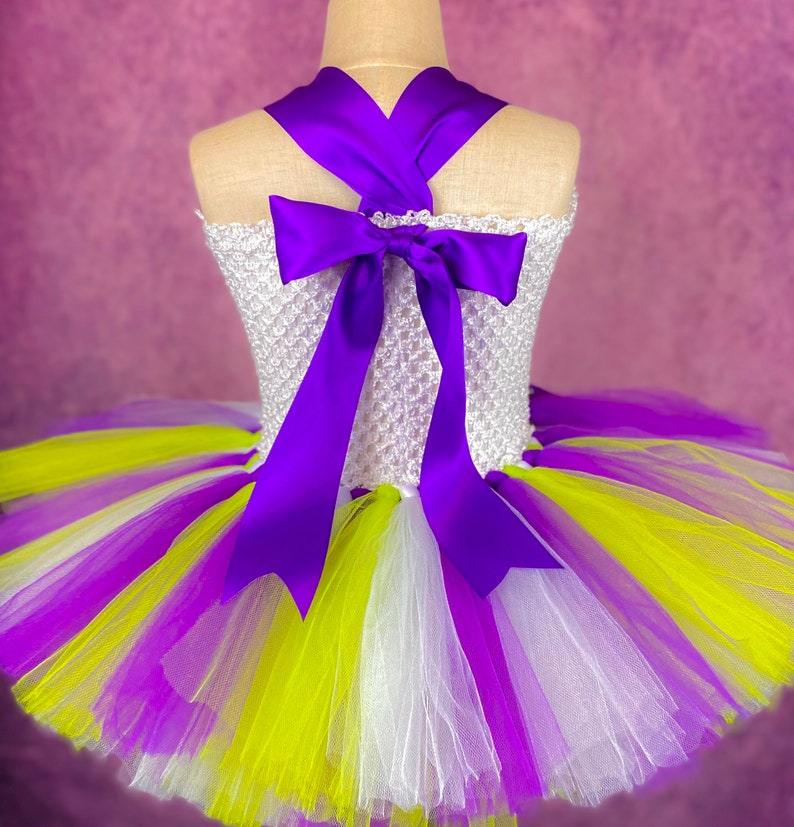 May include: A white tulle tutu skirt with purple, white, and yellow stripes. The skirt has a purple satin ribbon tied in a bow at the back.