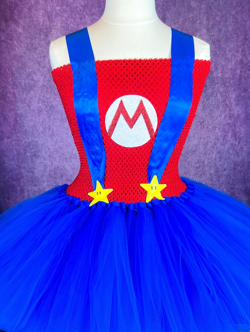 Pode incluir: Um tutu de tule azul com um corpete vermelho com um M branco com glitter e al&ccedil;as azuis. O corpete tem estrelas amarelas na parte inferior.