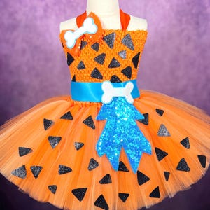 May include: An orange tutu dress with black triangle accents, a blue bow, and bone-shaped embellishments. The bodice is orange with black triangles, and the skirt is layered orange tulle. A blue ribbon and bone-shaped accents complete the look.
