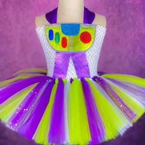 May include: A purple, white, and green tulle tutu with a white bodice featuring a purple bow and a colorful cartoon character design. The character has a yellow body, blue eyes, and red and green details.