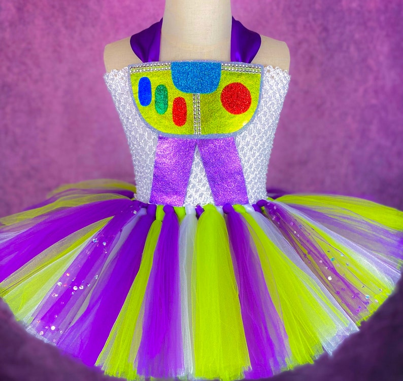 May include: A purple, white, and green tulle tutu with a white bodice featuring a purple bow and a colorful cartoon character design. The character has a yellow body, blue eyes, and red and green details.