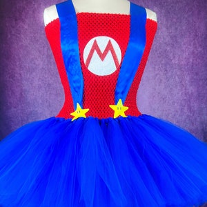 Pode incluir: Uma saia tutu de tule azul com um corpete vermelho e branco com um grande "M" e al&ccedil;as azuis. O corpete &eacute; decorado com duas estrelas amarelas.
