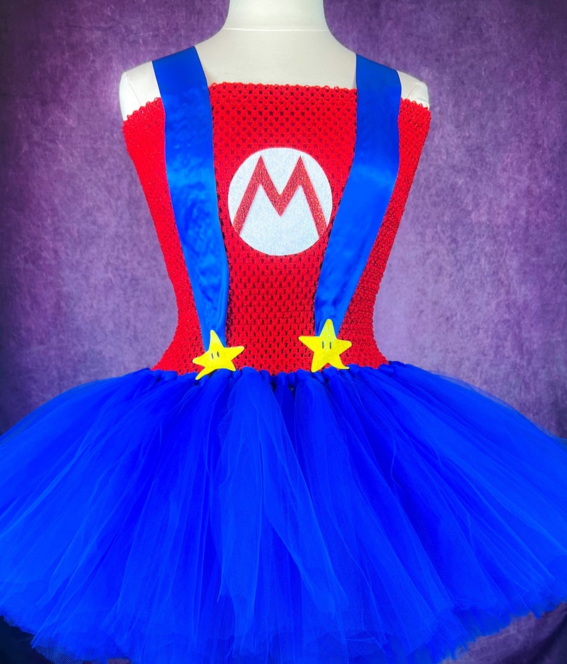 Pode incluir: Uma saia tutu de tule azul com um corpete vermelho e branco com um grande "M" e al&ccedil;as azuis. O corpete &eacute; decorado com duas estrelas amarelas.