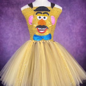 May include: A yellow tutu dress with a Mr. Potato Head design. The bodice is gold with a black hat, eyes, mustache, and a blue bow tie. The skirt is layered tulle with sparkling accents. The dress is displayed on a mannequin.