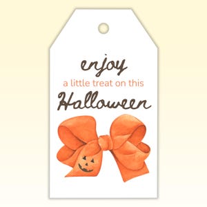 Puede incluir: Etiqueta de regalo blanca de Halloween con las palabras "enjoy a little treat on this Halloween" en escritura marrón y naranja. Un lazo naranja con una cara de calabaza está en la parte inferior.