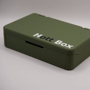Puede incluir: Una caja rectangular verde con un cajón y un asa de cuerda. La caja tiene el texto "Hott Box" impreso en la parte superior.