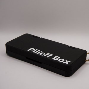 Pode incluir: Uma caixa retangular preta com um rótulo branco que diz "Piiiieff Box". A caixa tem um cordão marrom amarrado.