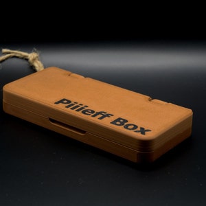 Puede incluir: Una pequeña caja de madera con un acabado marrón y un asa de cuerda. La caja tiene el texto "Piiiieff Box" grabado en la parte superior.