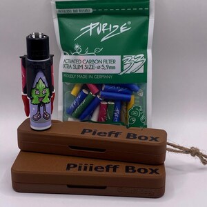 Puede incluir: Una caja de madera marrón con el texto "Pieff Box" y "Piiieff Box" en la parte superior e inferior de la caja. La caja está abierta y contiene un encendedor negro con un personaje de dibujos animados verde en él. Una bolsa de filtros de cigarrillos verdes, azules y rojos está al lado de la caja.