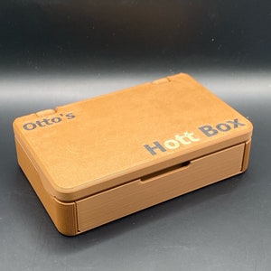 Może przedstawiać: Brązowe prostokątne pudełko z napisem "Otto's Hott Box" wydrukowanym na pokrywie. Pudełko ma wysuwaną szufladę i pokrywę na zawiasach. Pudełko jest wykonane z jasnobrązowego materiału.