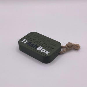 Könnte beinhalten: Eine kleine, rechteckige Schachtel in Dunkelgrün mit dem Aufdruck "TrAshBox" in weiß und schwarz. Die Schachtel hat eine Schnur.