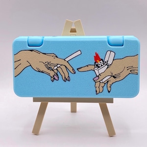 Könnte beinhalten: Ein hellblaues Etui mit einem Gemälde von zwei Händen, eine hält eine Zigarette und die andere ein Feuerzeug, auf einem kleinen Holzstaffelei.