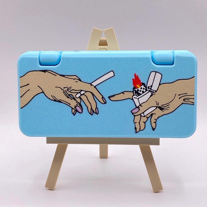 May include: A light blue case with a painting of two hands, one holding a cigarette and the other holding a lighter, on a small wooden easel.