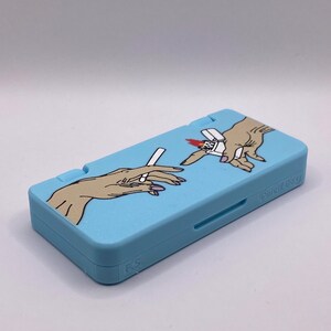 May include: A light blue Nintendo DS case with a graphic of two hands holding a cigarette and a lighter. The case has the text "Guilty Gear" printed on the side.
