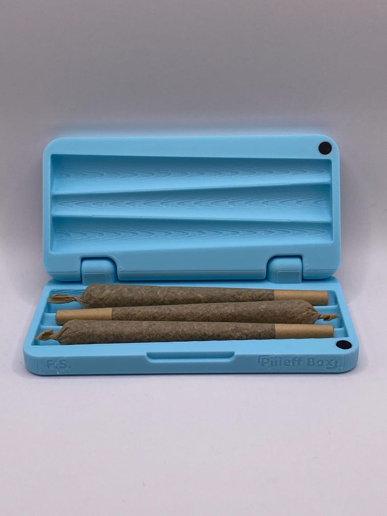 May include: A light blue, rectangular, plastic case with a black dot on the top right corner. The case is open and has four brown, rolled cigarettes inside. The case has the text "Pliff Box" on the bottom right corner and "FCS" on the bottom left corner.