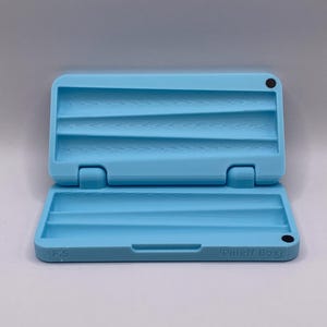 May include: A light blue, rectangular, plastic rolling tray with three slots. The tray is open and has the text "Puff Box" on the bottom.
