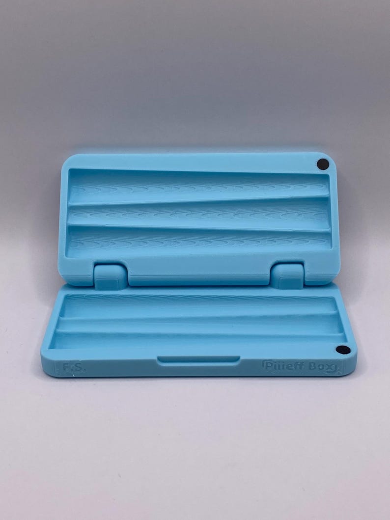 May include: A light blue, rectangular, plastic rolling tray with three slots. The tray is open and has the text "Puff Box" on the bottom.