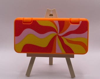 May include: Orange Nintendo Switch game case with a swirling yellow, pink, and red pattern. The case is on a small wooden easel.