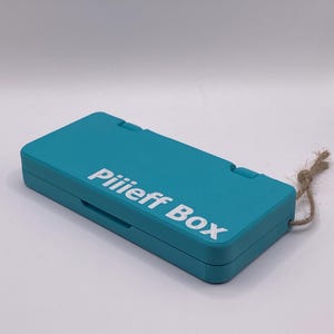 Może przedstawiać: Turkusowa plastikowa skrzynka z białym sznurkiem przymocowanym do górnej części. Na górze skrzynki widnieje napis "Pilieff Box" w kolorze białym.