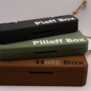 Puede incluir: Tres cajas de madera apiladas una encima de la otra. La caja superior es negra y dice "Pieff Box". La caja del medio es verde y dice "Pilieff Box". La caja inferior es marrón y dice "Hott Box".