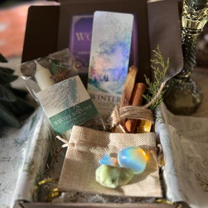 Puede incluir: Una caja de regalo con varios artículos, que incluyen una vela, anís estrellado, una tarjeta con una escena invernal, palitos de canela y piedras pequeñas. La caja está forrada con papel estampado y contiene una pequeña bolsa de yute.