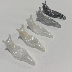 May include: A set of five small slug figurines arranged in a diagonal line. Each slug features a distinct glittery finish, ranging from a translucent white at the front to a dark charcoal gray at the back, with varying levels of sparkle.