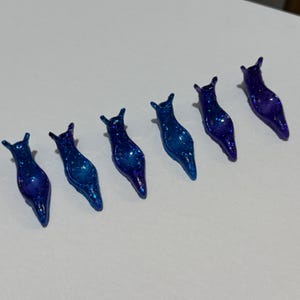May include: Six blue glitter slug-shaped resin charms. The charms are various shades of blue, with a glittery, sparkling effect. Each charm has a small, pointed head and a curved body. They are arranged in a diagonal line.