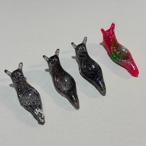 May include: Four glittery slug figurines. Three are dark with silver glitter, and one is red with green and silver glitter. The slugs have small, pointed antennae and are arranged on a white surface.