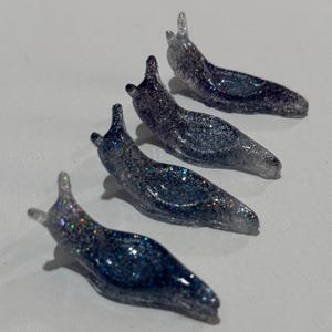 May include: Four small slug-shaped figurines made of a translucent, dark blue and black resin material. Each figurine is embedded with fine, multi-colored glitter that catches the light, creating a sparkling effect across the smooth surfaces.