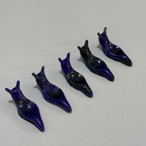 May include: Five small, slug-shaped figurines arranged in a diagonal line. Each figurine features a glossy, deep purple finish with subtle dark, shimmering glitter accents integrated into the surface, creating a smooth, reflective appearance.