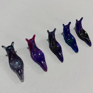 May include: Five small slug figurines arranged in a row. Each slug features a glossy, semi-translucent finish with embedded glitter particles. The colors range from deep purple and blue to magenta, with varying levels of sparkle and iridescent shimmer.