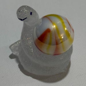 May include: A small glass snail figurine with a translucent, glittery, textured body and a smooth, spherical shell. The shell is white with swirling orange and yellow stripes. The snail has a simple dark blue painted eye and mouth.
