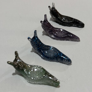 May include: Four slug-shaped figurines arranged in a diagonal line. Each figurine features a distinct glittery finish in shades of silver, blue, purple, and black. The objects have smooth, curved bodies with two small tentacles at the head.