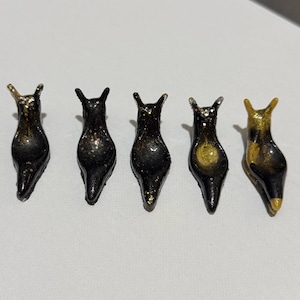 May include: Five handmade slug figurines. The slugs are black with gold and silver glitter accents. Each slug has a unique color pattern, with some featuring gold or silver highlights on their backs and antennae. The figurines are displayed on a white surface.