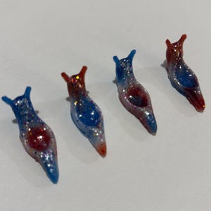 May include: Four small slug-shaped figurines arranged in a row. Each figurine features a translucent gradient of red and blue colors with embedded glitter particles throughout the body and two small antennae on the head.