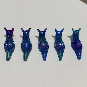 May include: Five glittery, slug-shaped resin figurines. Each slug is a different shade of blue, green, and purple, with a sparkly, iridescent finish. The figurines are arranged in a row, showcasing their unique color variations.