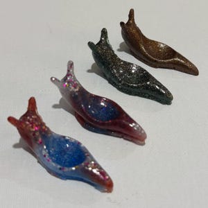 May include: A set of three slug-shaped figurines with hollowed-out centers. One is blue and red with multi-colored glitter, one is dark gray with silver glitter, and one is a solid metallic bronze color with a subtle shimmer finish.