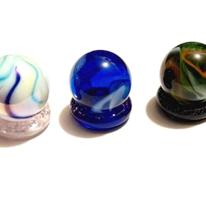以下が含まれることがあります： ディスプレイスタンドに置かれたガラス製のビー玉3つ。ビー玉は、白の渦巻き模様が入った青色、青と紫の渦巻き模様が入った白色、オレンジと白の渦巻き模様が入った緑色です。