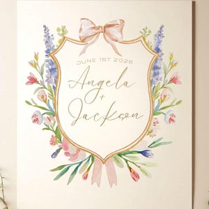 Op de afbeelding: Waterverf trouwkaart met een goudomrand schild met de namen "Angela + Jackson" en de datum "1 juni 2026". Het schild is versierd met een perzikkleurige strik en omgeven door kleurrijke bloemen en groen.