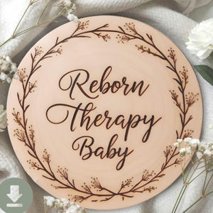 May include: A round wooden sign with the words "Reborn Therapy Baby" in elegant script. The sign is encircled by a delicate, brown floral wreath. The background is a soft, neutral color, with white flowers and fabric.