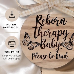 May include: A round wooden ornament featuring the black text Reborn Therapy Baby. Please be kind. with a black butterfly illustration on each side. The ornament is topped with a beige and white checkered ribbon. Digital download and print icons are visible.