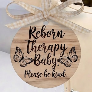 Puede incluir: Un letrero redondo de madera con la frase "Reborn therapy Baby. Please be kind." en escritura negra, con ilustraciones de mariposas. Una cinta de cuadros beige y blanca está atada en la parte superior.