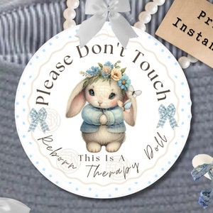 以下が含まれることがあります： ウサギのイラストと「Please Don't Touch Reborn Doll This Is A Therapy」というテキストが入った丸いサイン。タグには「Print Instantly」と書かれています。ウサギは青いセーターと花の冠をかぶっています。