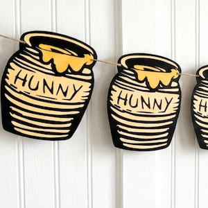 May include: A decorative banner featuring multiple honey pot cutouts. Each pot is black with yellow honey spilling over the top and the word "HUNNY" in the center. The banner is strung on a natural twine.
