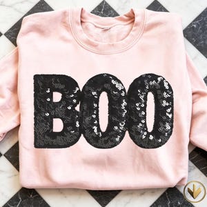 May include: A light pink crewneck sweatshirt featuring the word BOO centred on the front in large, bold, black sequin lettering. The garment has long sleeves, a ribbed neckline, and a soft fabric texture, showcasing a clear, high-contrast design.